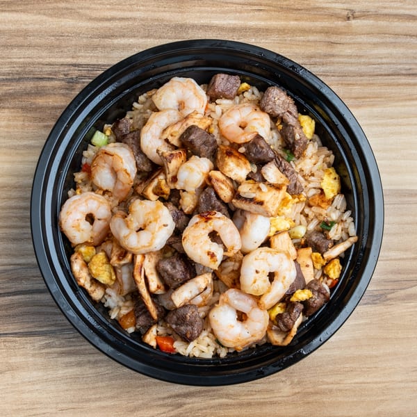 Overhead view of a black bowl filled with fried rice, shrimp, steak, and chicken on a wooden table.