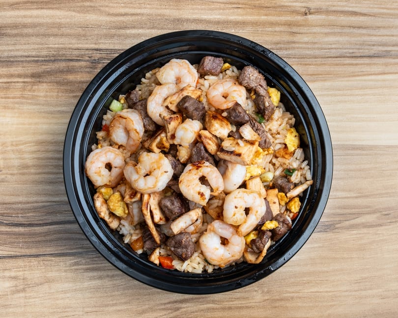 Overhead view of a black bowl filled with fried rice, shrimp, steak, and chicken on a wooden table.