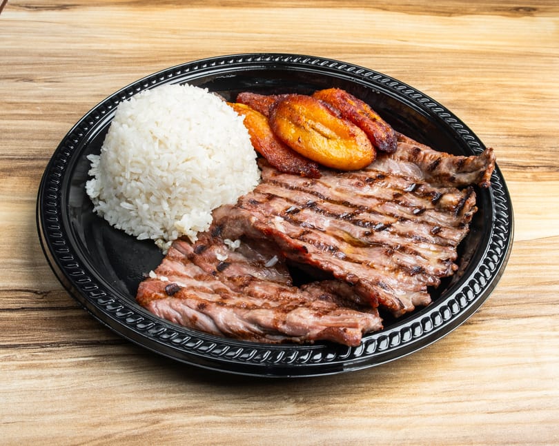 Grilled steak, white rice, and fried plantains served on a black plate on a wooden table.