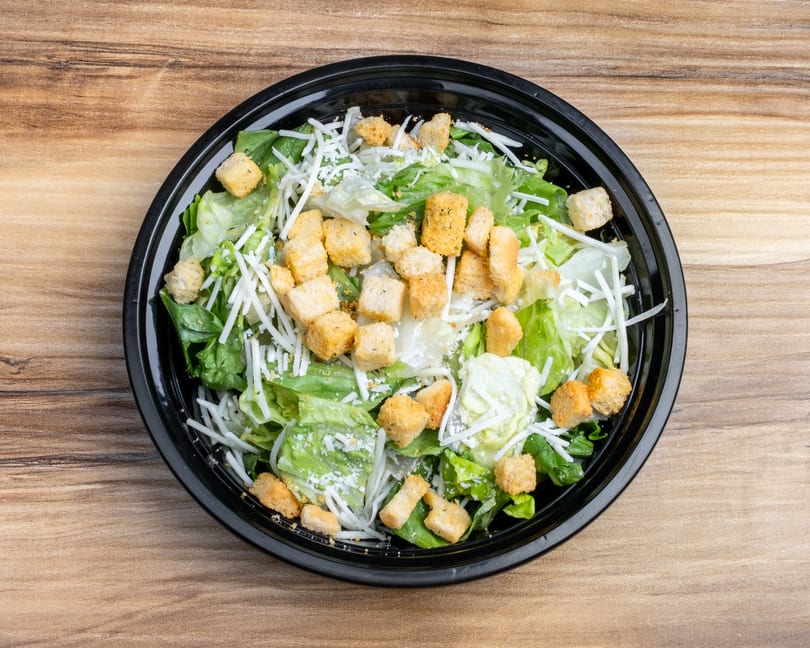 Overhead view of a Caesar salad with lettuce, croutons, and shredded cheese in a black bowl.