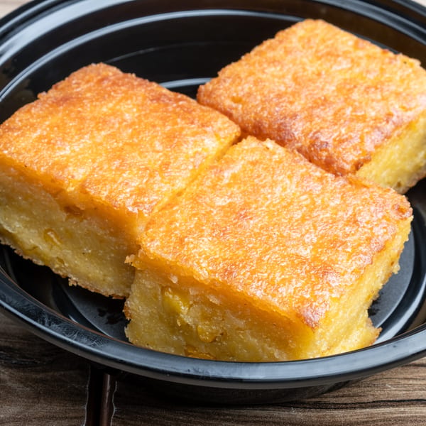 Three golden-brown squares of baked cassava cake with a slightly crispy top, in a black bowl.