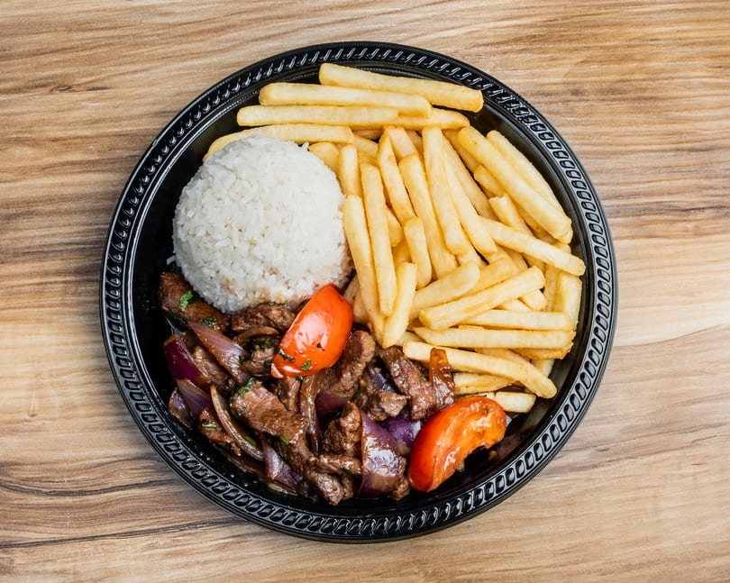 Lomo Saltado with steak, red onions, tomatoes, white rice, and french fries on a black plate.